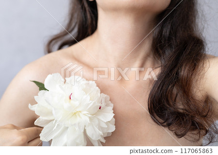 a young woman brunette holds a white peony flower near her tongue bare shoulder, beauty concept 117065863