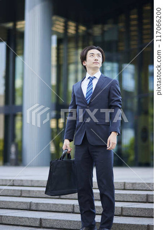 A young businessman in a suit looking into the distance and an office building A young businessman in a suit looking into the distance and an office building 117066200