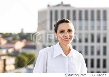 Smiling woman in white shirt standing outdoors in urban setting Smiling woman in white shirt standing outdoors in urban setting 117066544