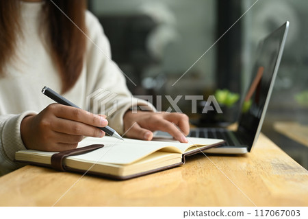 Cropped shot of young woman taking notes on notebook and using laptop at a coffee shop 117067003