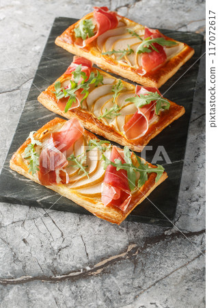 Delicious baked puff pastries with pear, cream cheese, jamon and arugula close-up on marble board. Vertical 117072617