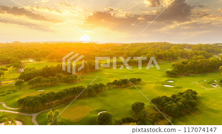Golf course at sunset with beautiful sky and sand trap. Scenic panoramic view of golf fairway with bunker. Golf field with pines Golf course at sunset with beautiful sky and sand trap. Scenic panoramic view of golf fairway with bunker. Golf field with pines 117074147