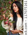 Gorgeous brunette female with long hair, picking apples in apple orchard. Portrait of Caucasian beauty wearing white chemise posing in garden with closed eyes in evening. Femininity, harvest concept. 117075161