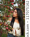 Pensive brunette with bright necklace, picking apples in orchard in fall. Portrait of Caucasian beauty wearing white chemise posing in garden with closed eyes at day. Concept of femininity, harvest. 117075162