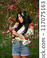 Thoughtful, serious woman in jeans shorts, touching apple tree branches, laden with fruits. Front view of young woman with long dark hair posing in apple orchard with closed eyes. Harvesting concept. 117075163
