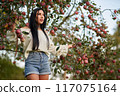 Long haired brunette walking through apple orchard, looking ahead. Low angle view of nice adult girl in casual apparel, posing in apple garden in early autumn, blurred background. Harvest concept.  117075164