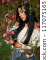 Serious young woman thinking, while leaning on apple tree branches at warm autumn day. Front view of calm Caucasian beauty with dark hair in apple orchard. Concept of emotions, female beauty, fall. 117075165
