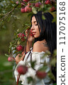 Gorgeous relaxed woman leaning on apple tree branch in apple orchard. View from shoulder of tattooed beauty wearing white shoulderless blouse in garden at twilight. Concept of female beauty, harvest. 117075168