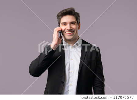 Adult 30s businessman speaks with smile on mobile phone. Guy holding and using smartphone. Formal suit outfit. Grey studio background Adult 30s businessman speaks with smile on mobile phone. Guy holding and using smartphone. Formal suit outfit. Grey studio background 117075573
