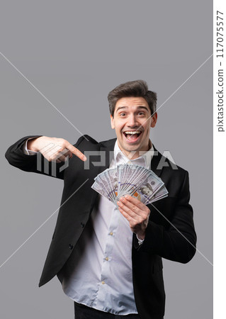 Happy excited businessman with cash money - USD currency dollars banknotes on grey wall. Symbol of jackpot, gain, victory, winning Happy excited businessman with cash money - USD currency dollars banknotes on grey wall. Symbol of jackpot, gain, victory, winning 117075577