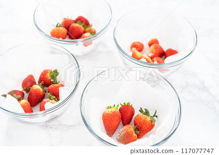 Bright red strawberries, interspersed with signs of mold, rest in a glass bowl lined with a paper towel on a white napkin, indicating improper storage techniques. 117077747