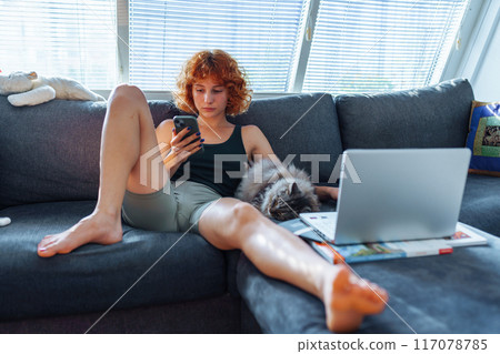 Portrait of red-haired young woman sitting on sofa using laptop, grey cat sleeping nearby 117078785