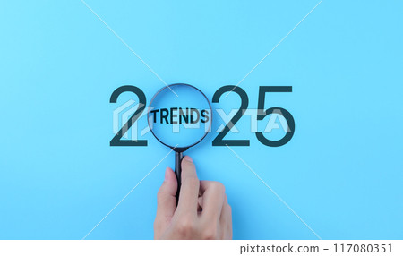 Business Trends 2025 Concept. A hand holds a magnifying glass focusing on the word "Trends" within the year "2025," representing the concept of future business and marketing trends and planning. 117080351