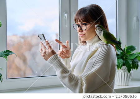 Middle aged woman and parrot together at home using smartphone, near winter window 117080636