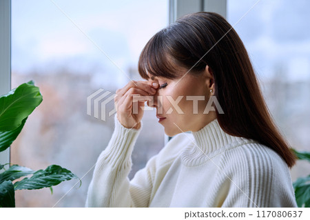 Middle aged woman experiencing headache migraine, at home near window Middle aged woman experiencing headache migraine, at home near window 117080637