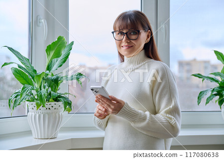 Smiling middle aged woman holding smartphone, at home near window Smiling middle aged woman holding smartphone, at home near window 117080638
