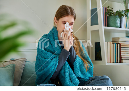 Young sick woman sitting under warm blanket sneezing coughing in handkerchief 117080643