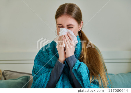 Young sick woman sitting under warm blanket sneezing coughing in handkerchief 117080644