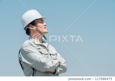 一名身穿工作服的亞洲男工戴著頭盔,仰望藍天下的天空 一名身穿工作服的亞洲男工戴著頭盔,仰望藍天下的天空 117086475