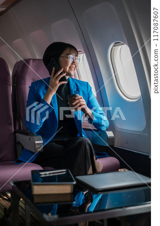 Professional woman in blue blazer using smartphone and drinking coffee on airplane. Business travel concept. 117087607