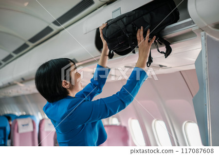 Flight attendant in blue uniform placing a bag in the overhead compartment inside an airplane cabin. Flight attendant in blue uniform placing a bag in the overhead compartment inside an airplane cabin. 117087608