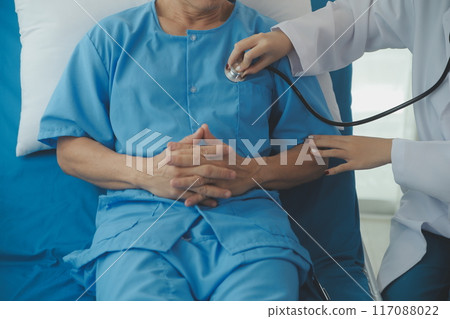 Doctor using stethoscope to listen senior man heart during examination in hospital room and wearing visor as safety precaution against coronavirus. Medical control for infections, disease. 117088022