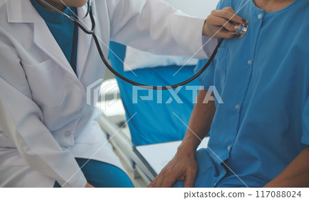 Doctor using stethoscope to listen senior man heart during examination in hospital room and wearing visor as safety precaution against coronavirus. Medical control for infections, disease. 117088024