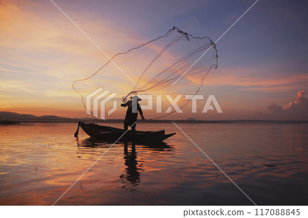 Fisherman casting his net on during sunset. Silhouette Asian fisherman on wooden boat casting a net for freshwater fish. Thailand culture. Fisherman casting his net on during sunset. Silhouette Asian fisherman on wooden boat casting a net for freshwater fish. Thailand culture. 117088845