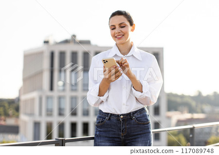 Smiling woman using smartphone on rooftop with city view 117089748