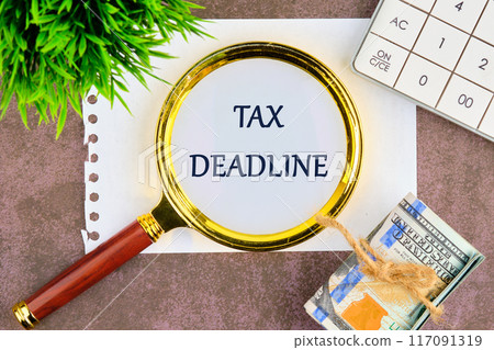 TAX DEADLINE words written through a magnifying glass on a piece of paper in a composition with banknotes, a plant and a calculator 117091319