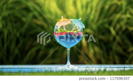 Glass with a blue cocktail, garnished with fruit and mini umbrellas, on the pool edge with green grass in the background 117096307