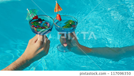 Two hands holding cocktail glasses against the water surface of a pool, refreshing drinks with fruit and soda 117096350