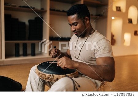 Calm African man in trance sitting playing on steel tongue drum with sticks on floor in meditation room. Drummer using traditional meditative instrument glucophone. Concept of mindfulness, tranquility 117097022