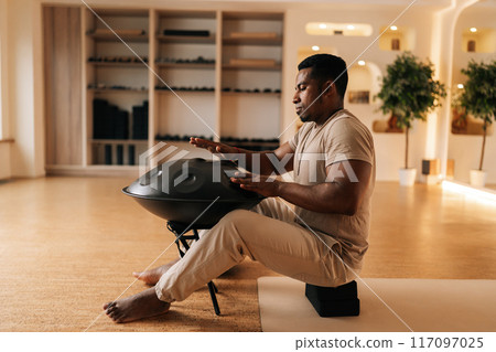 Relaxed African young man in trance sitting playing on steel tongue drum by hands on floor at home with cozy interior. Silence calm filling room by sounds of music from musical instrument. 117097025