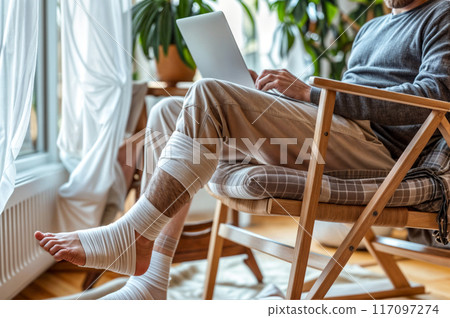 Man with broken leg sitting in chair using laptop computer. 117097274