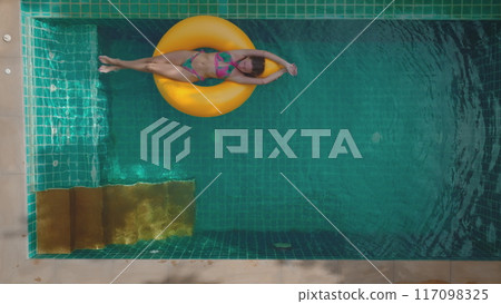 Young woman is enjoying her vacation, floating on yellow inflatable ring in swimming pool. Concept of summer vacation, relaxing in resort 117098325