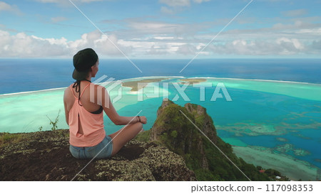 Young woman relaxes and meditates on a cliff edge, enjoying the breathtaking panoramic view of the turquoise lagoon and lush islands of maupiti in french polynesia Young woman relaxes and meditates on a cliff edge, enjoying the breathtaking panoramic view of the turquoise lagoon and lush islands of maupiti in french polynesia 117098353