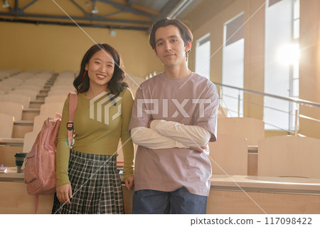 Medium full shot of two students of Asian and mixed background leaning on desk in empty sunlit lecture hall, while smiling and looking at camera at university Medium full shot of two students of Asian and mixed background leaning on desk in empty sunlit lecture hall, while smiling and looking at camera at university 117098422