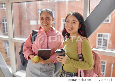 Portrait shot of two cheerful multiethnic smiling female students on snack break looking at camera while standing in university hallway next to window 117098441