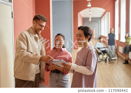 Two happy students casually talking to smiling adult male professor asking him for advice in pink university hallway, copy space Two happy students casually talking to smiling adult male professor asking him for advice in pink university hallway, copy space 117098442