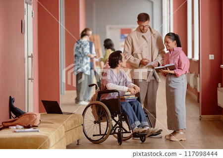 Adult male professor answering question in friendly informal conversation with male student using wheelchair and smiling female learner in pink university hallway 117098444