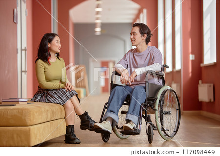 Two young multiethnic students chatting away amicably in university hallway. Smiling young man with disability talking to Asian girl, copy space Two young multiethnic students chatting away amicably in university hallway. Smiling young man with disability talking to Asian girl, copy space 117098449