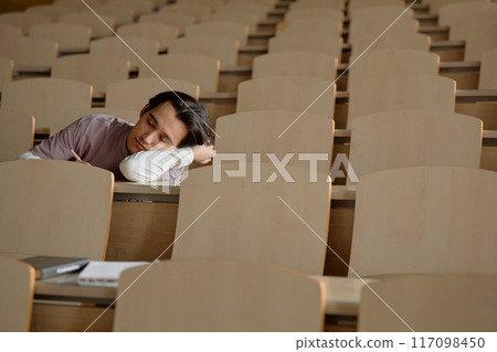 Wide angle shot of tired male student sleeping on desk while studying alone in lecture hall, copy space Wide angle shot of tired male student sleeping on desk while studying alone in lecture hall, copy space 117098450