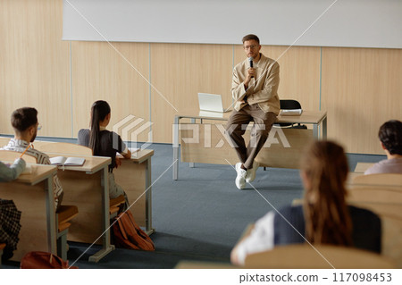 Adult male teacher with hand held microphone lecturing university students while leaning on desk in lecture hall, copy space Adult male teacher with hand held microphone lecturing university students while leaning on desk in lecture hall, copy space 117098453