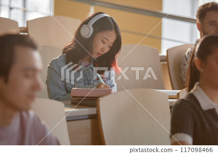 Disinterested Asian female student in headphones listening to music while drawing in notebook in lecture hall at university, copy space 117098466