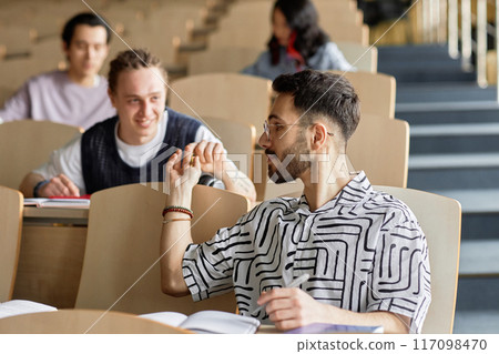 Side view shot of young male Middle Eastern student at desk secretly passing classmate behind him note while attending class in lecture hall at university Side view shot of young male Middle Eastern student at desk secretly passing classmate behind him note while attending class in lecture hall at university 117098470