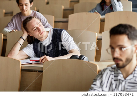 Bored young dreadlocked male student with head leaning on hand attending lecture at university Bored young dreadlocked male student with head leaning on hand attending lecture at university 117098471