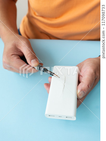 Close up of hands repairing white power bank with screwdriver on blue background. Close up of hands repairing white power bank with screwdriver on blue background. 117098733