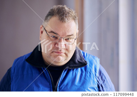 Large, plump-cheeked man of Caucasian ethnic group in prescription glasses has frowning look from under his eyebrows. 117099448