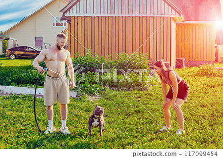 Young owners of an American Bully dog are having fun with their pet in backyard of country house, dog drinks water from garden hose. Young owners of an American Bully dog are having fun with their pet in backyard of country house, dog drinks water from garden hose. 117099454